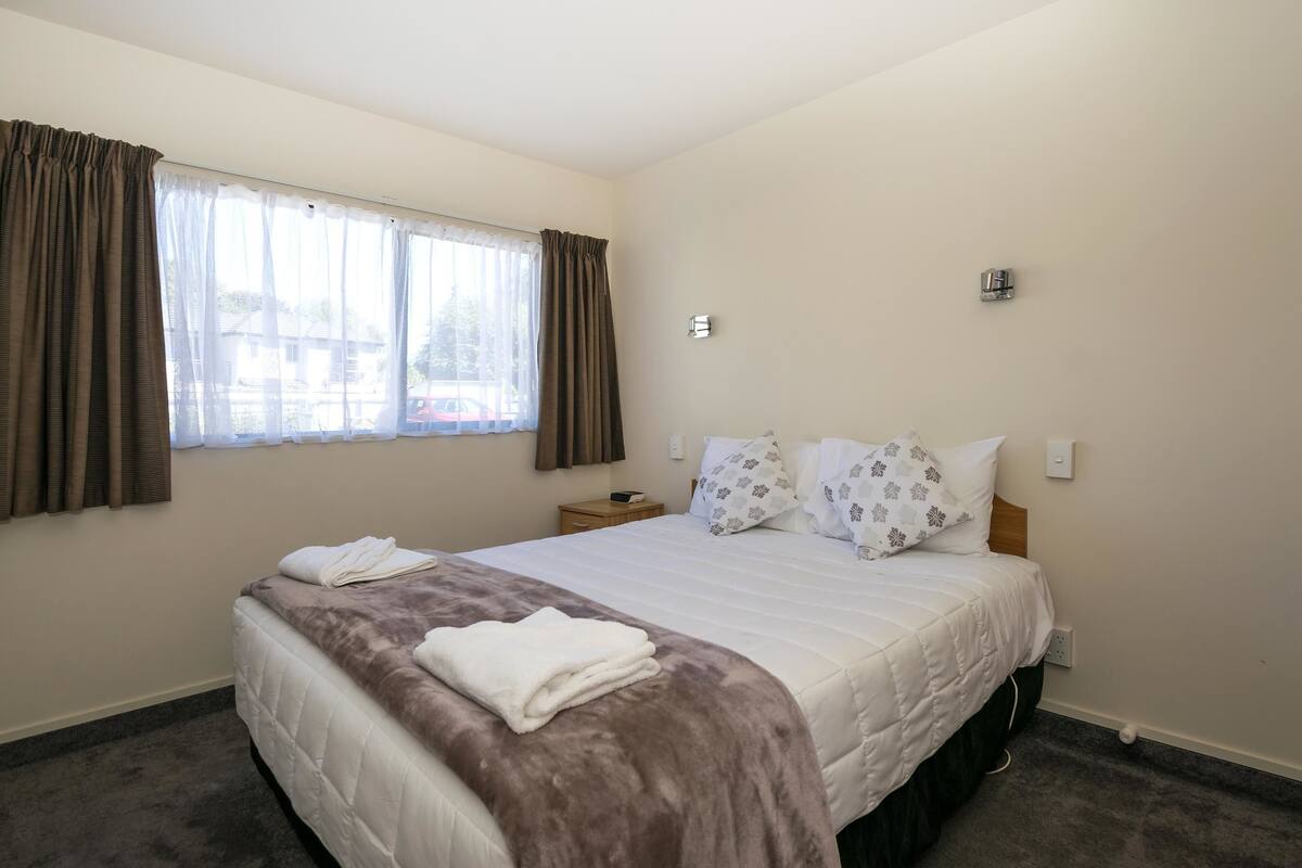 A hotel room with a large bed, a nightstand, a window with curtains, and a wall-mounted light.