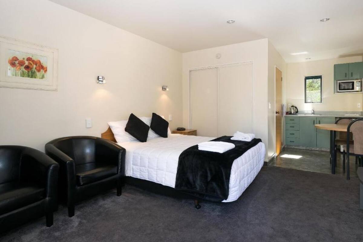 A bedroom with a bed, two black chairs, a nightstand, a painting, a closet, and a kitchenette with a microwave.