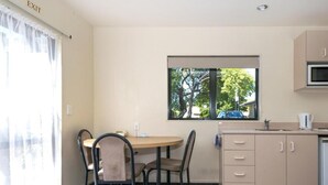 A small, well-lit room with a table and chairs, a window with a view of trees, and a kitchenette with cabinets and a microwave. - Ashleigh Court Motel (Blenheim)