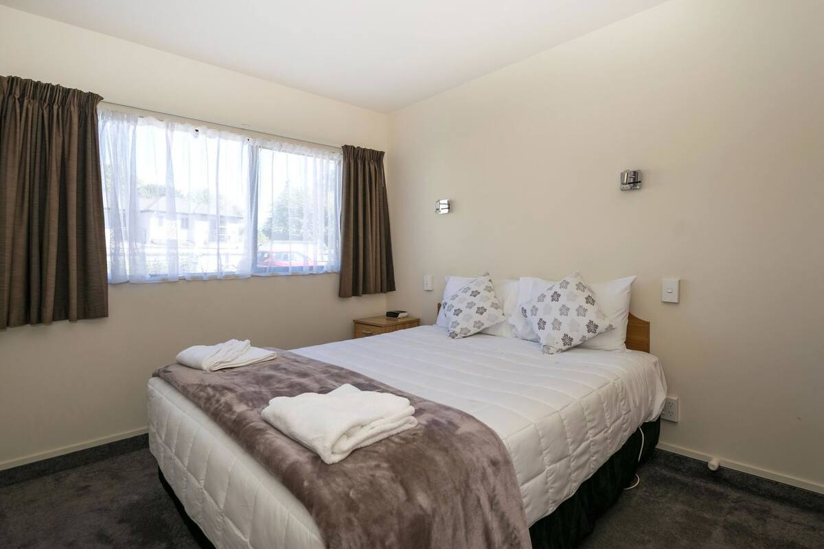 A hotel room with a bed, a nightstand, a window with curtains, and a wall-mounted light.