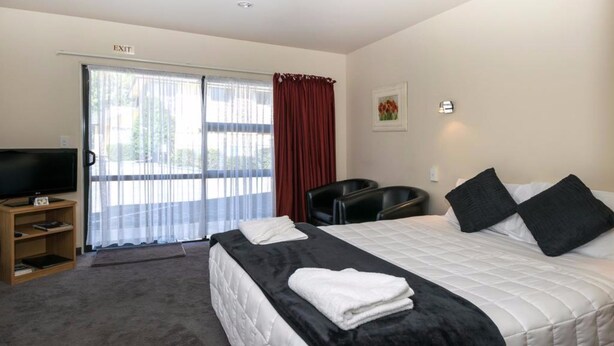 A hotel room with a bed, a TV, a desk with a chair, and a sliding glass door to the outside. - Ashleigh Court Motel (Blenheim)