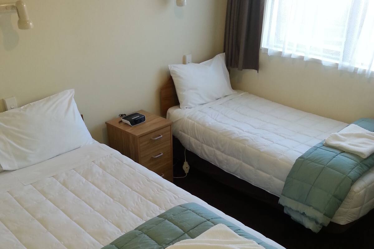 A hotel room with two beds, a nightstand, a lamp, and a window with curtains.
