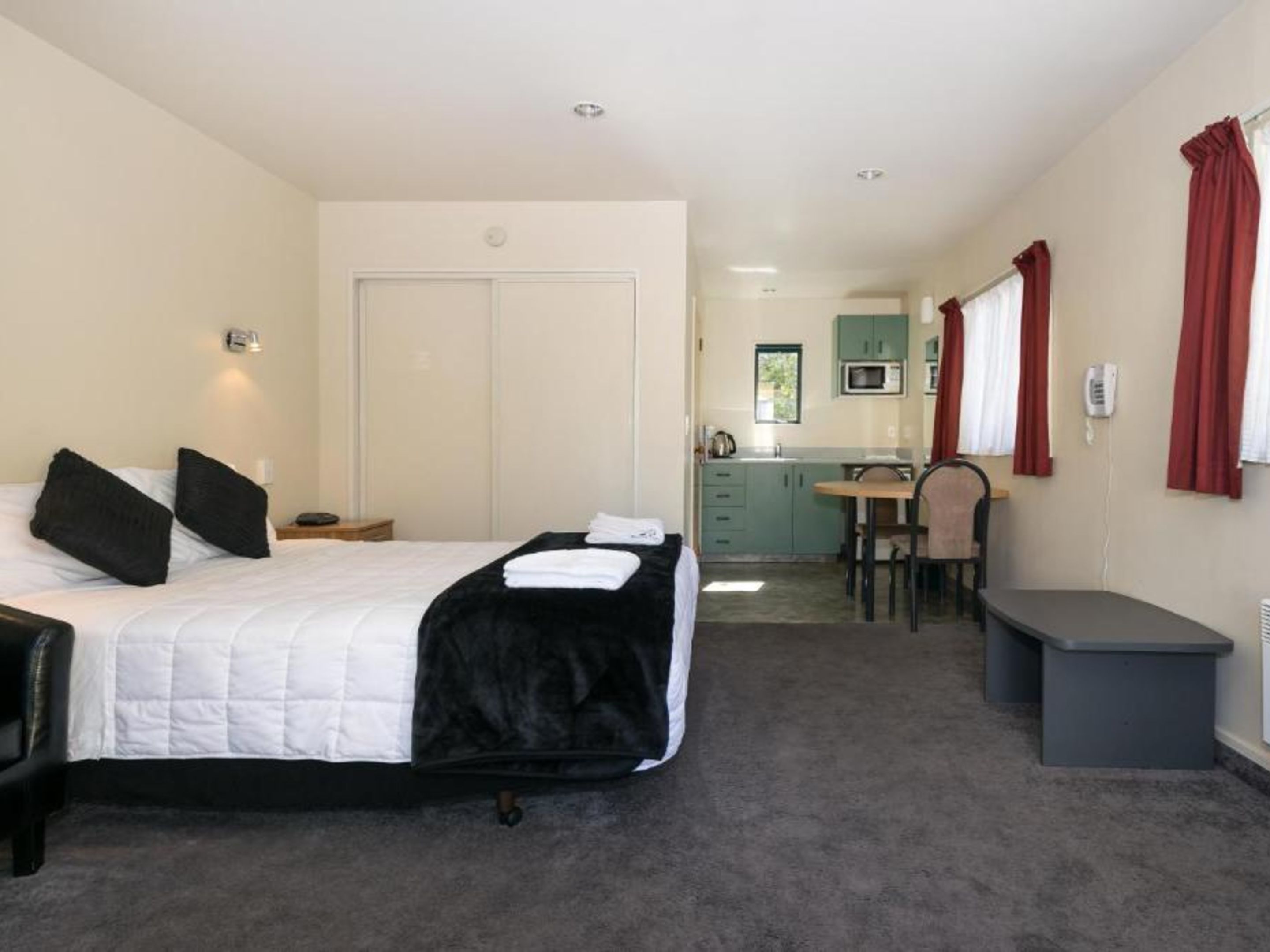 A hotel room with a bed, a desk, a chair, a small table, and a kitchenette in the background.