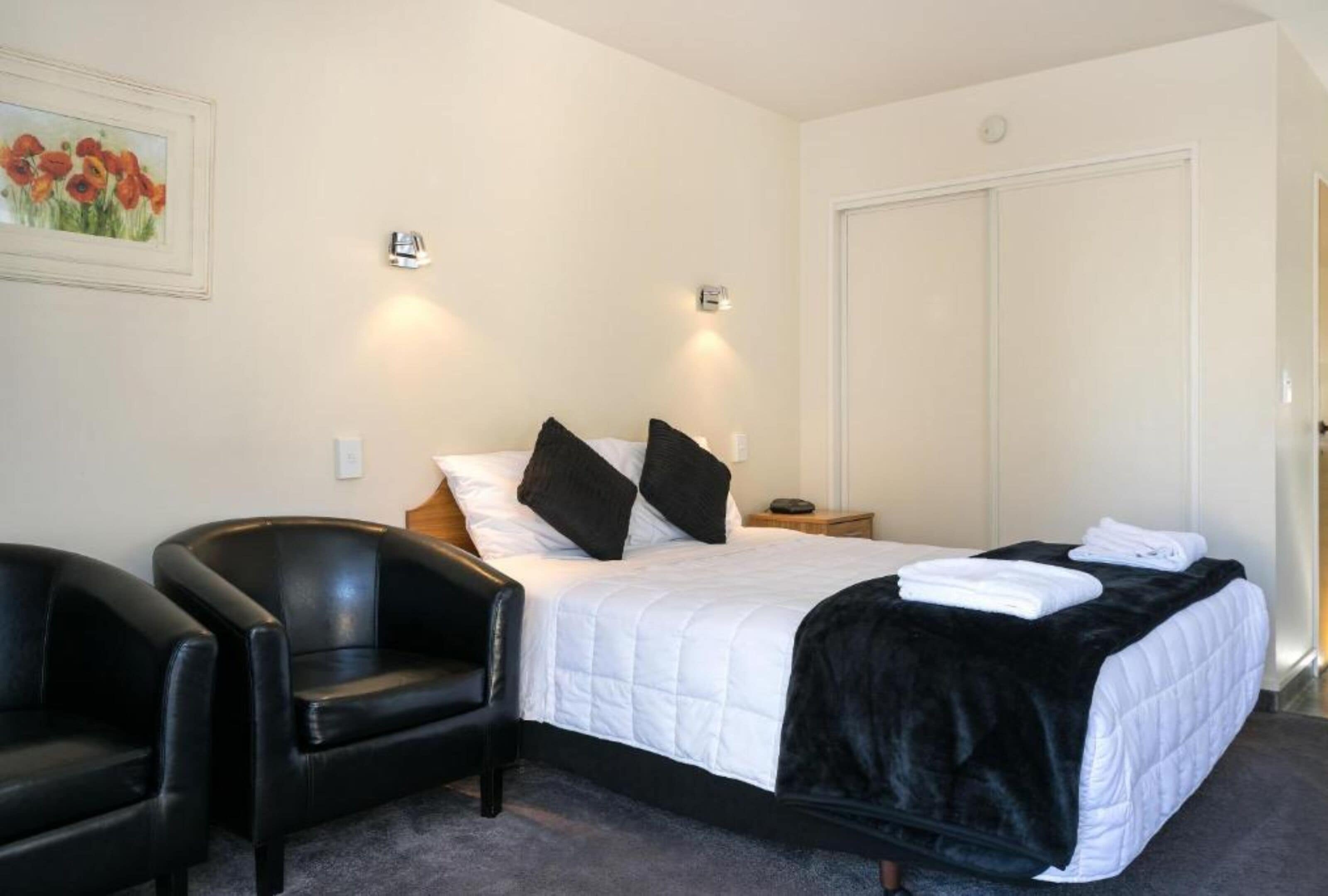 A neatly arranged bedroom with a bed, two bedside tables, a black leather chair, and a painting on the wall.