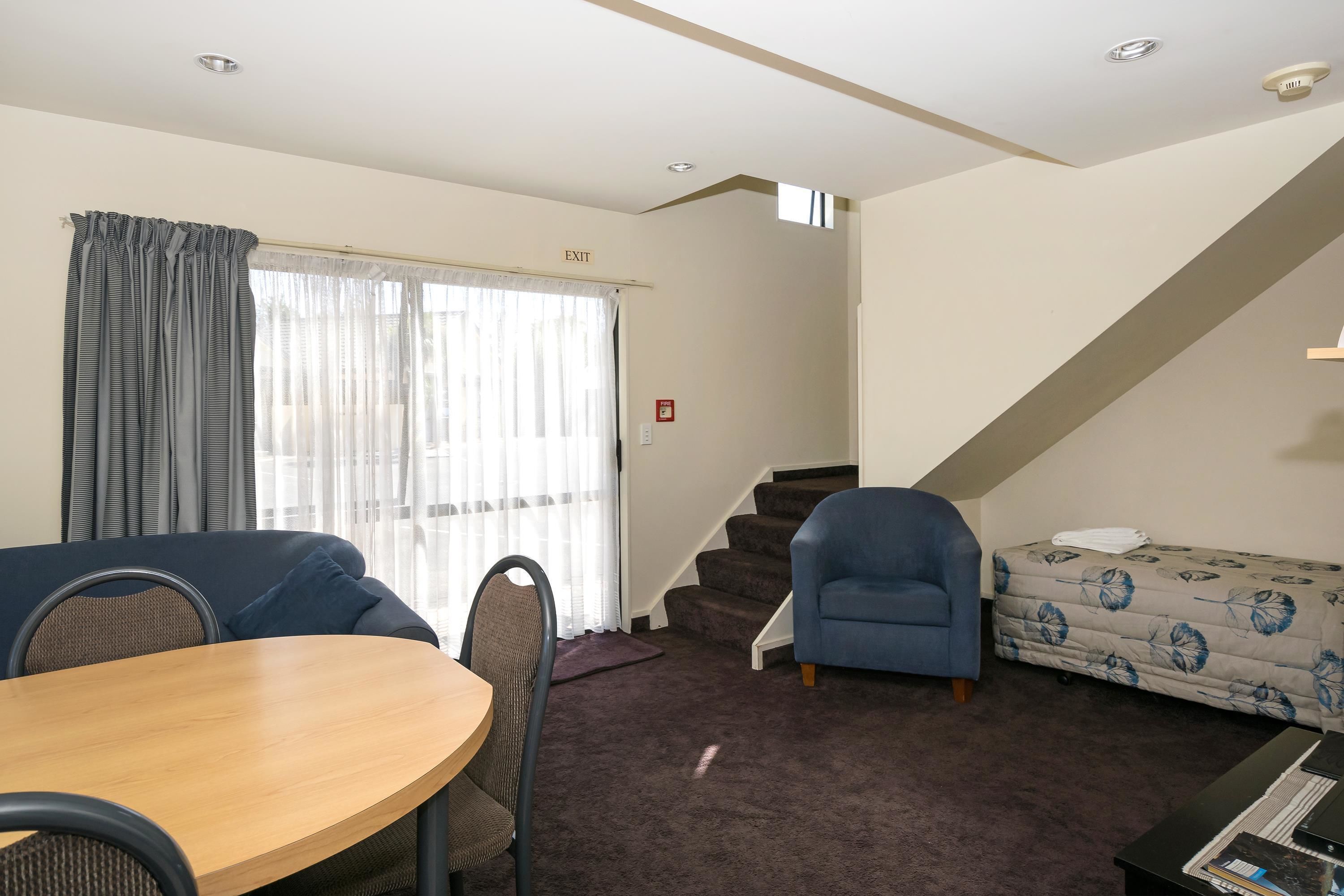 A room with a dining table, chairs, a sofa, a blue armchair, a bed with a floral duvet, a staircase, and a window with curtains.
