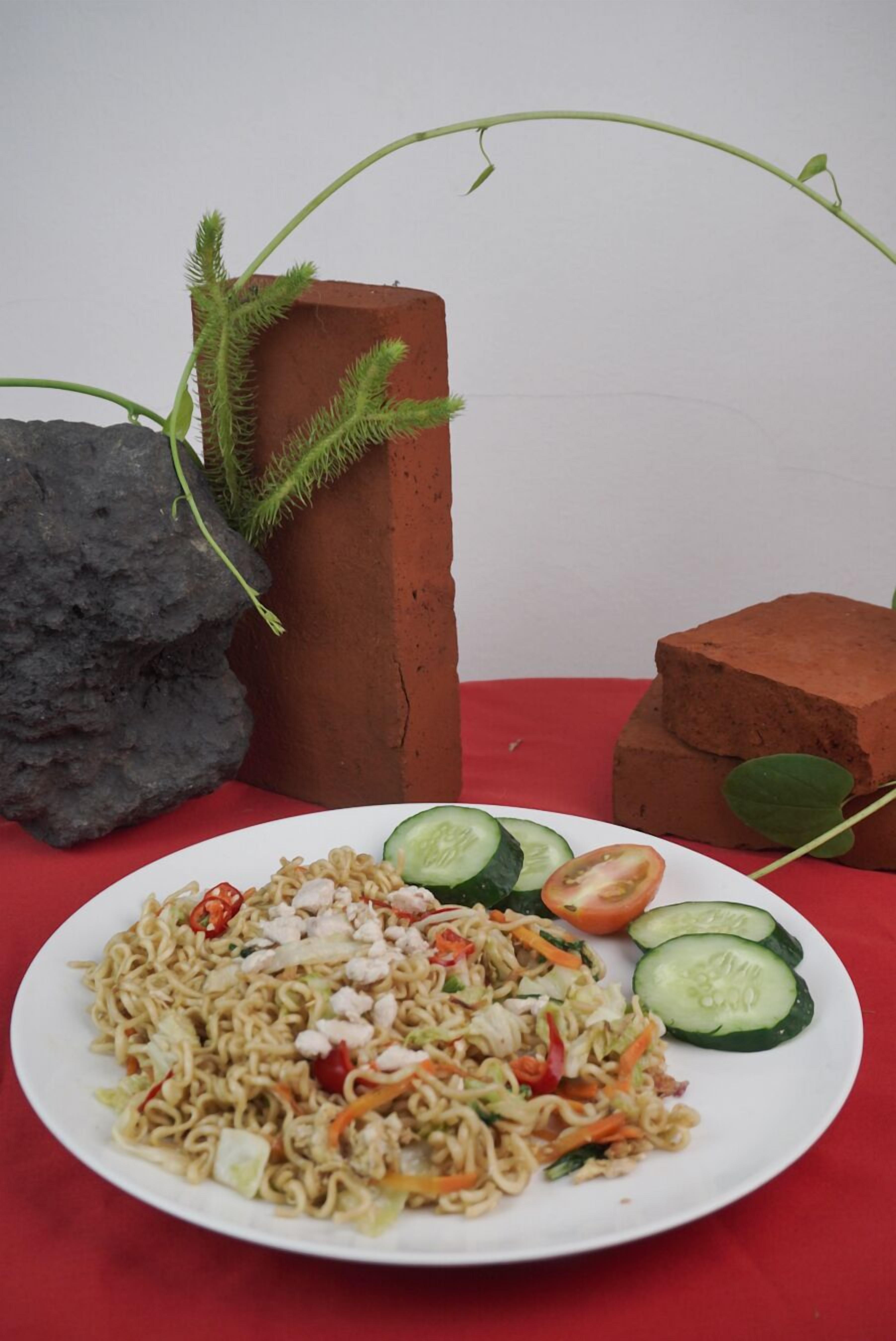 A plate of noodles with vegetables and meat, accompanied by cucumber slices on a red tablecloth.