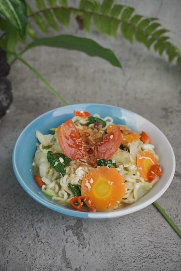 A bowl of noodles with vegetables and a slice of tomato on a blue plate. - Temple Cafe & Seaside Cottages (Karangasem)