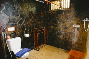Un baño con pared de piedra, inodoro, cabina de ducha de bambú y ventana con barrotes.