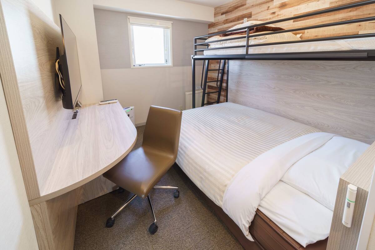 A bunk bed with a wooden headboard, a desk with a chair, a window, and a wall-mounted TV.