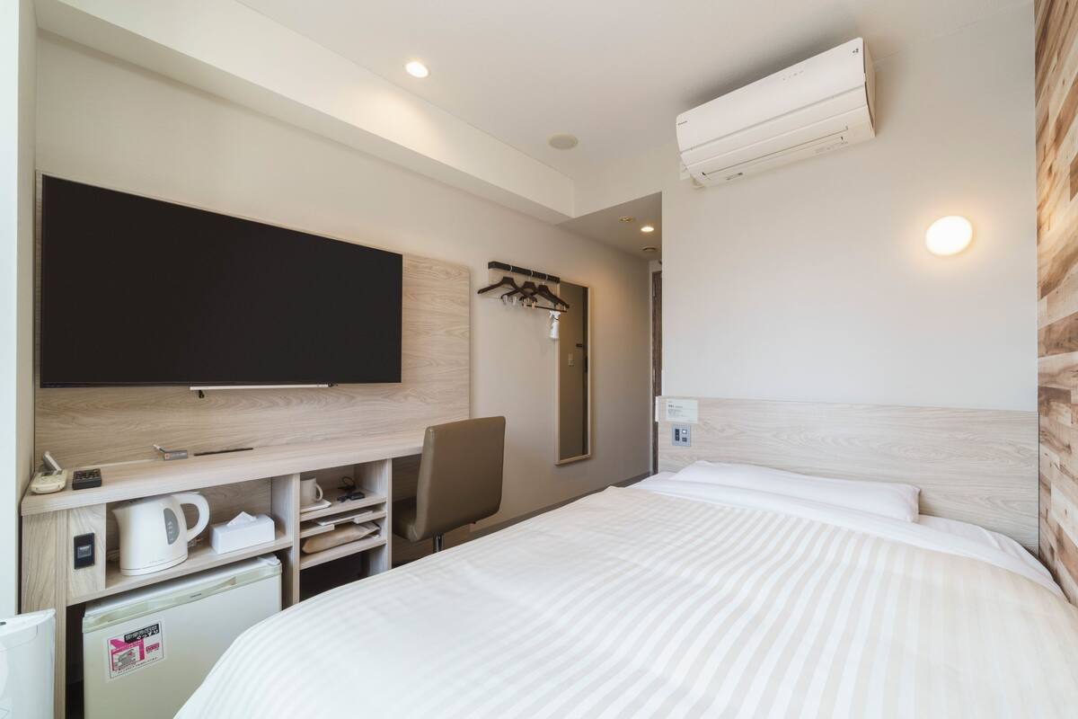 A hotel room with a bed, a TV mounted on the wall, a desk with a kettle, a small fridge, and a wooden headboard.