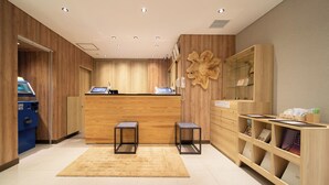 A hotel reception area with a wooden reception desk, two black chairs, a blue self-service machine, and a wooden cabinet with glass doors. - SUPER HOTEL Kushiro Kurogane (Kushiro)