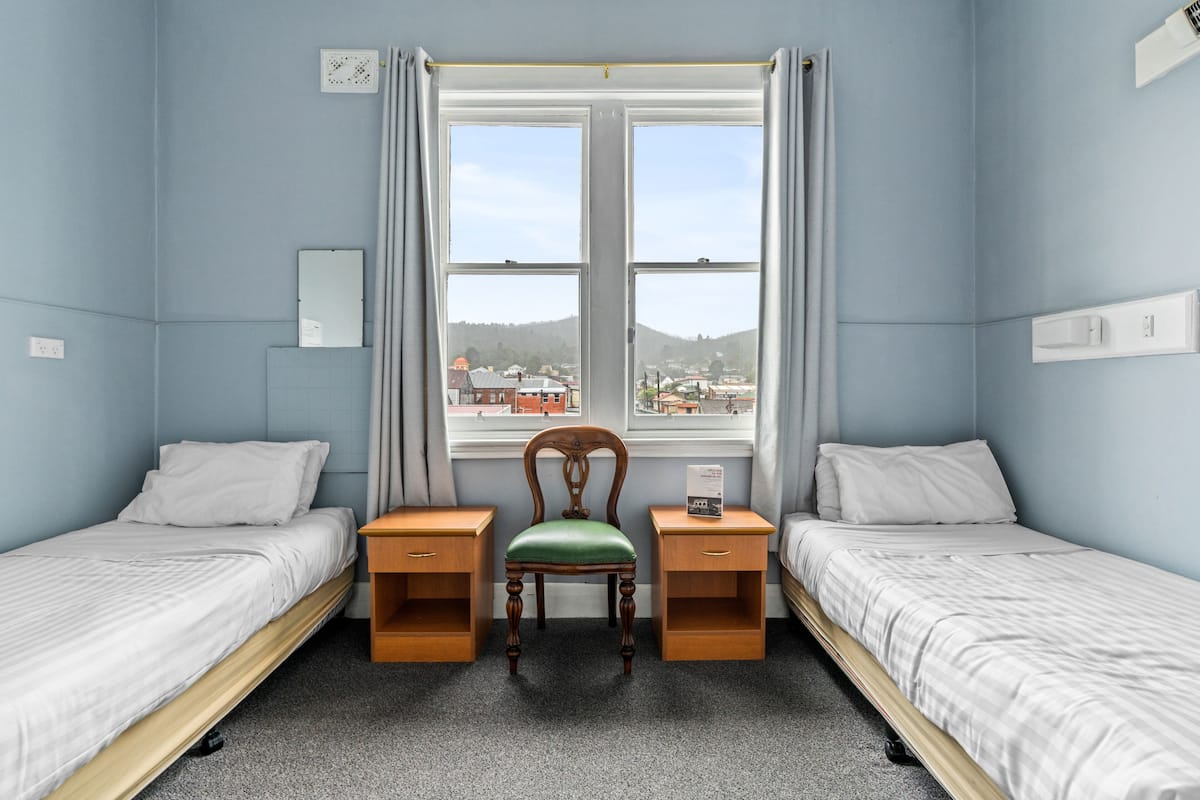 Ein Zimmer mit zwei Einzelbetten, einem Holzstuhl, einem Schreibtisch und einem Fenster mit Blick auf Gebäude.