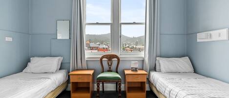 Ett rum med två separata sängar, en trästol, ett skrivbord och ett fönster med utsikt över byggnader.