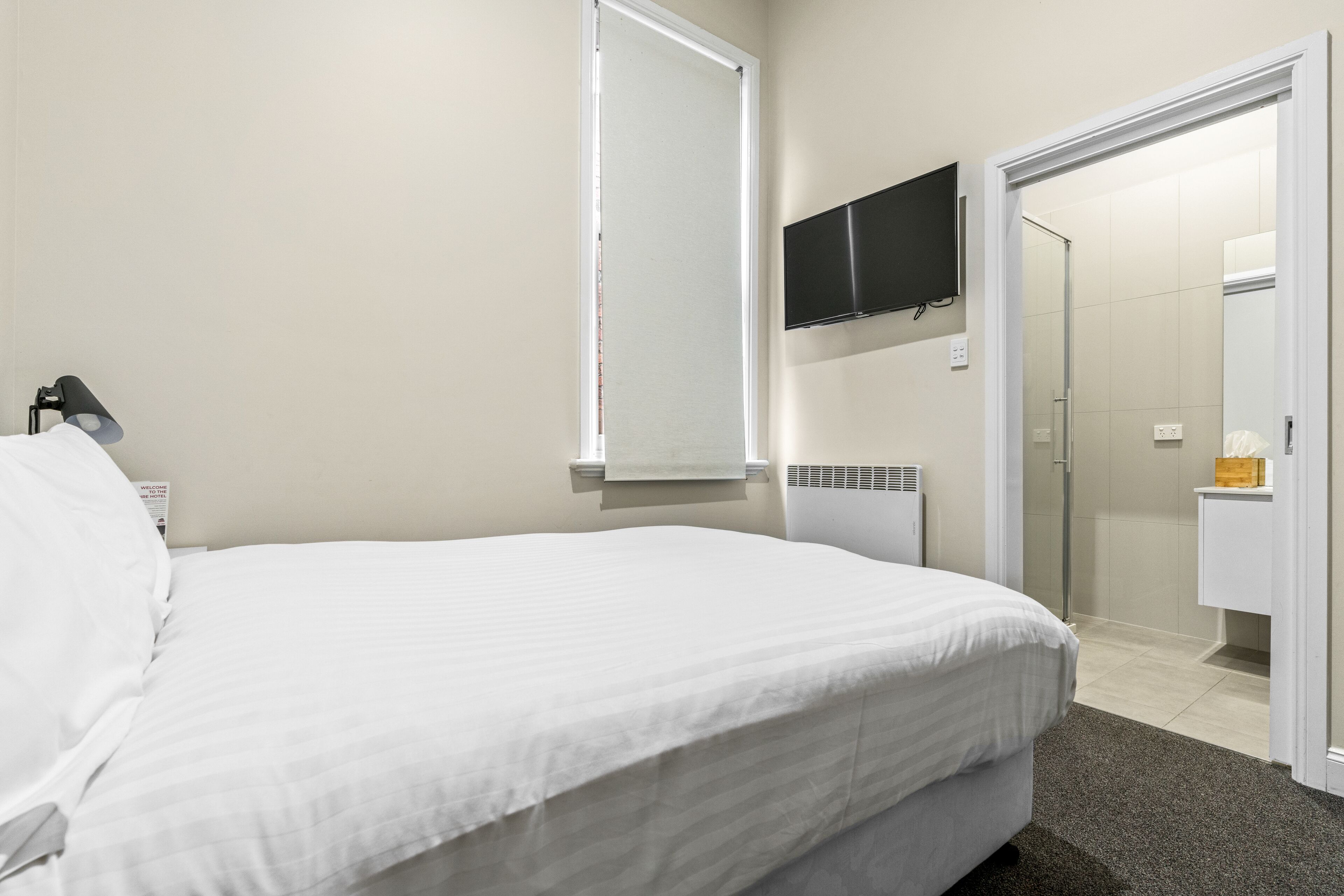 Una habitación de hotel con una cama, televisión montada en la pared, una ventana y una puerta que da a un baño.