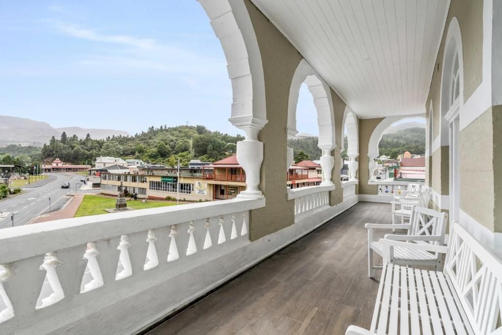 A balcony with white railings and benches, overlooking a street with buildings and a hilly landscape.