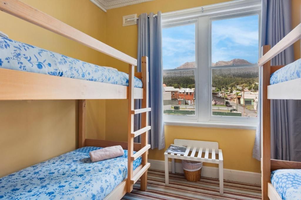 A room with bunk beds, a window with a view of buildings and mountains, a small table, and a chair.