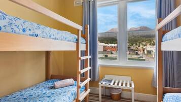 A room with bunk beds, a window with a view of buildings and mountains, a small table, and a chair.