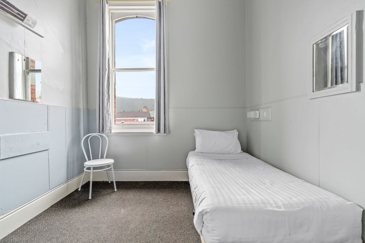 Ein schmaler, einfach eingerichteter Raum mit einem Bett, einem Stuhl, einem Fenster und einer Heizung.