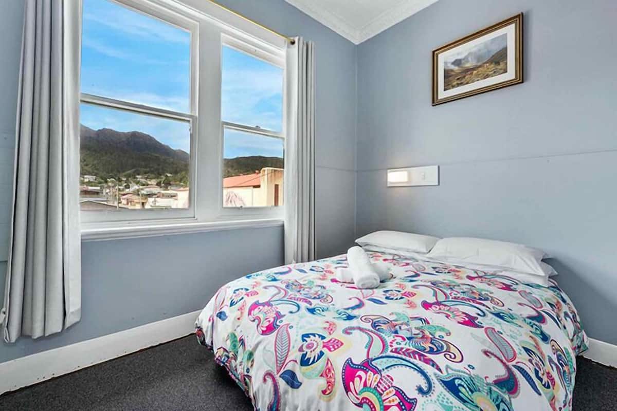 Ein Schlafzimmer mit Bett, einem fensterartigen Ausblick mit Vorhängen, einem Gemälde an der Wand und einer Aussicht auf eine Stadt und Berge.