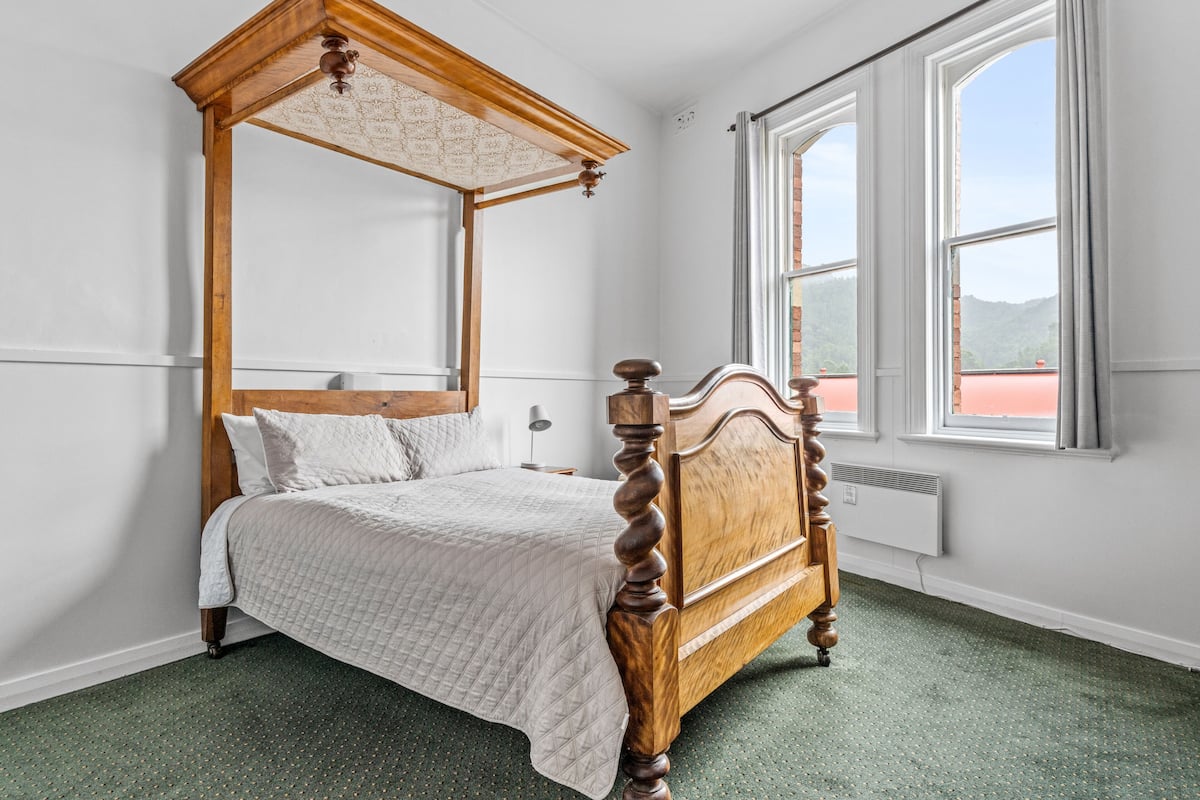 Ein Himmelbett mit Holzrahmen und Baldachin, vor einer weißen Wand mit einem großen Fenster platziert.