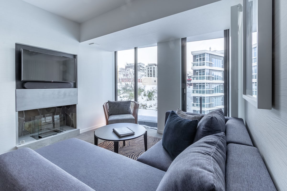 Una sala de estar moderna con un sofá gris, una mesa redonda de centro, una chimenea y un ventanal con vistas a la ciudad.