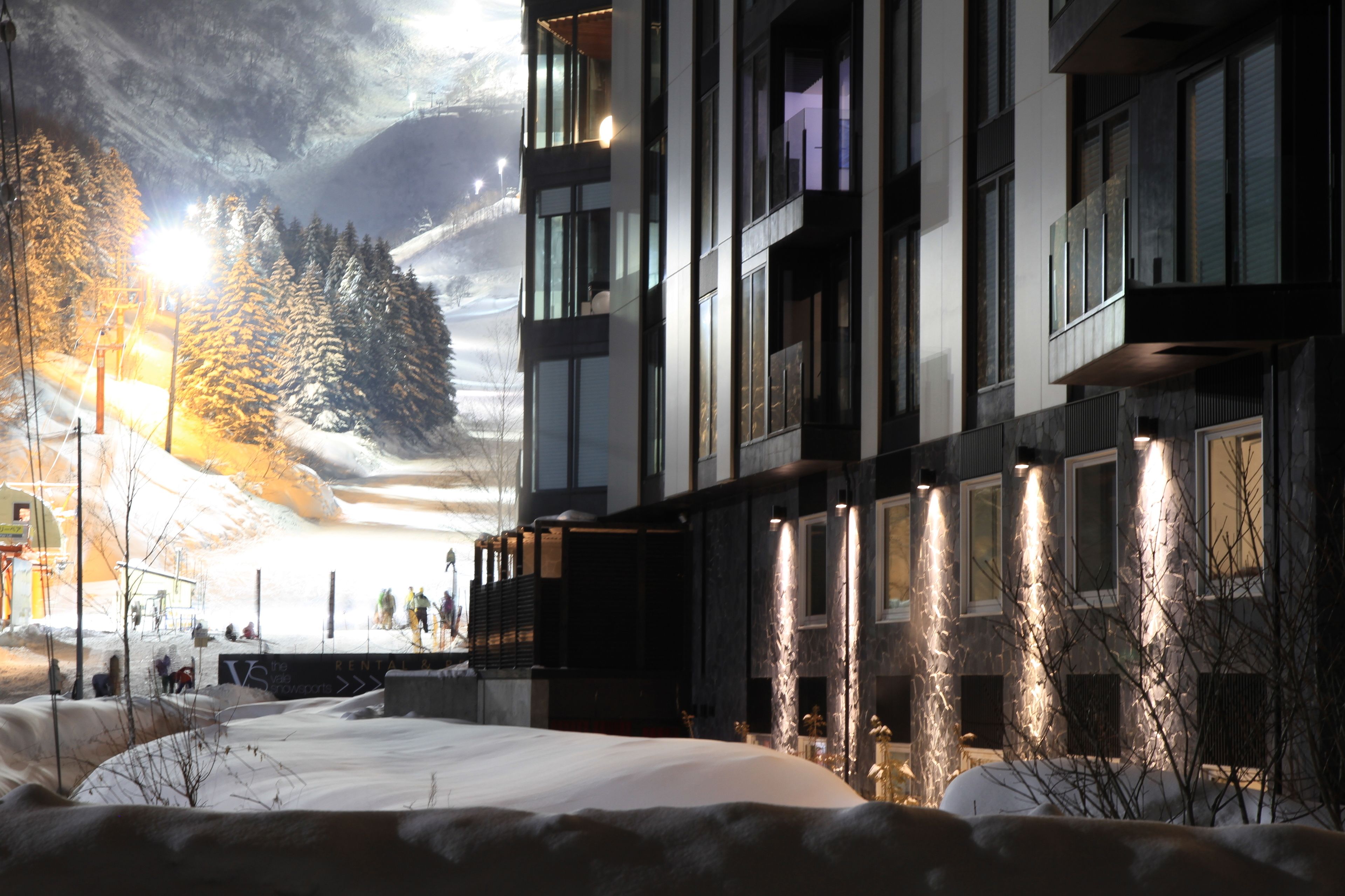 Un edificio moderno con varios balcones, suelo cubierto de nieve y montañas al fondo.