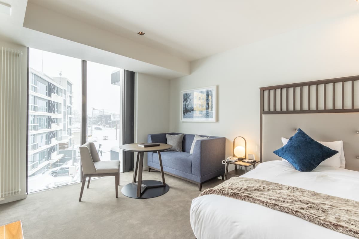 Una habitación de hotel moderna con un ventanal grande, un sofá, una mesa redonda, una cama y una mesita de noche.