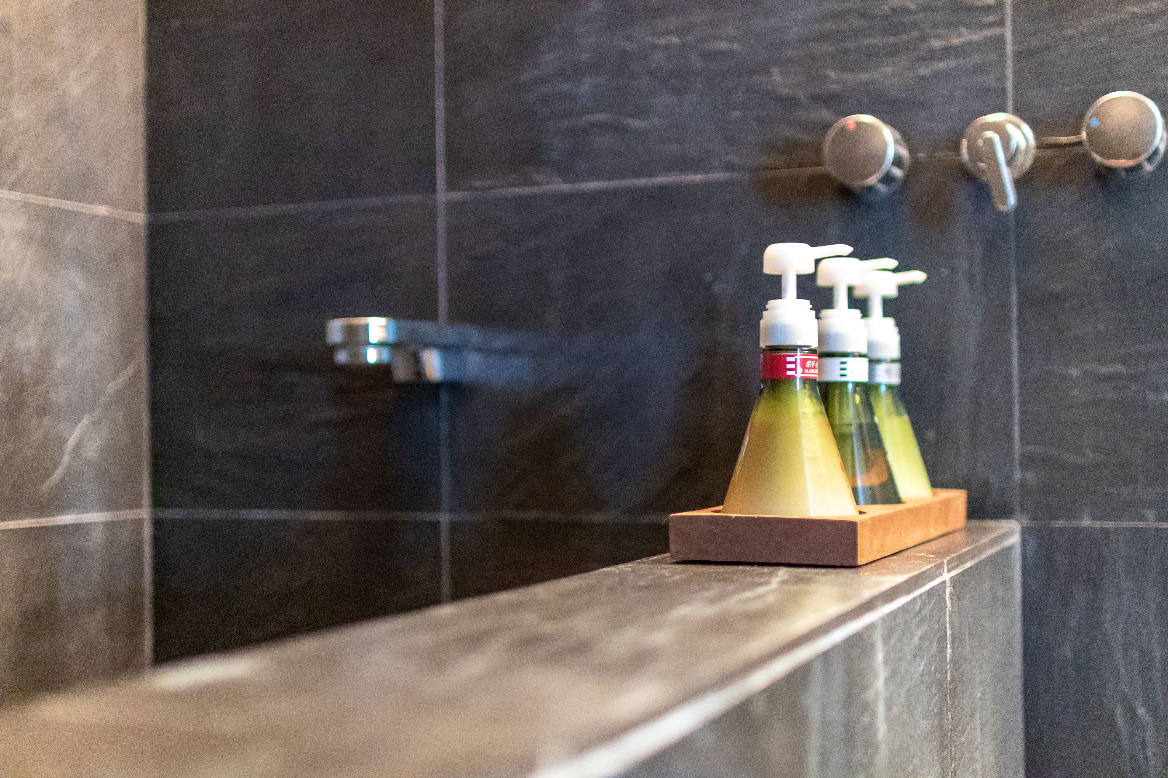 Un baño con baldosas oscuras, un porta jabón de madera y un cabezal de ducha.