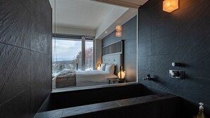 A modern bathroom with a large bathtub, a visible window, and a bed in the background. - The Vale Niseko (Kutchan)