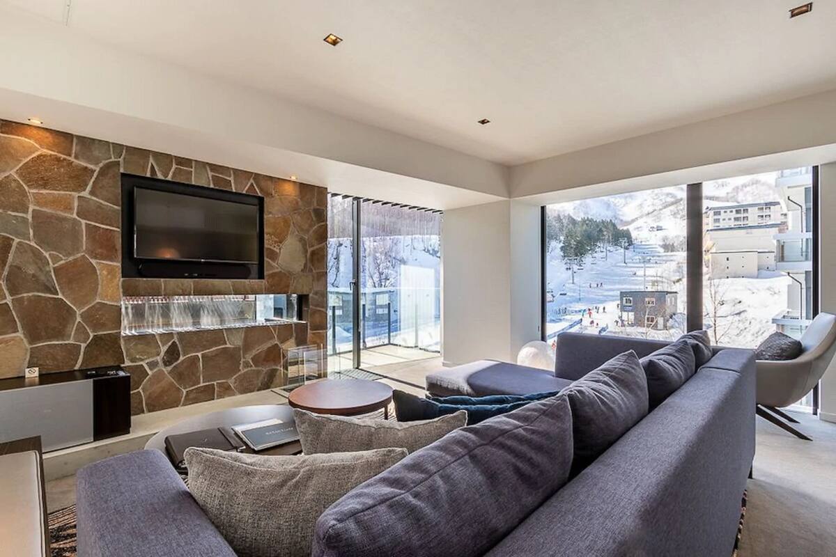 Una sala de estar moderna con una pared de piedra, un televisor de pantalla plana, un sofá y un amplio ventanal con vistas a un paisaje nevado.