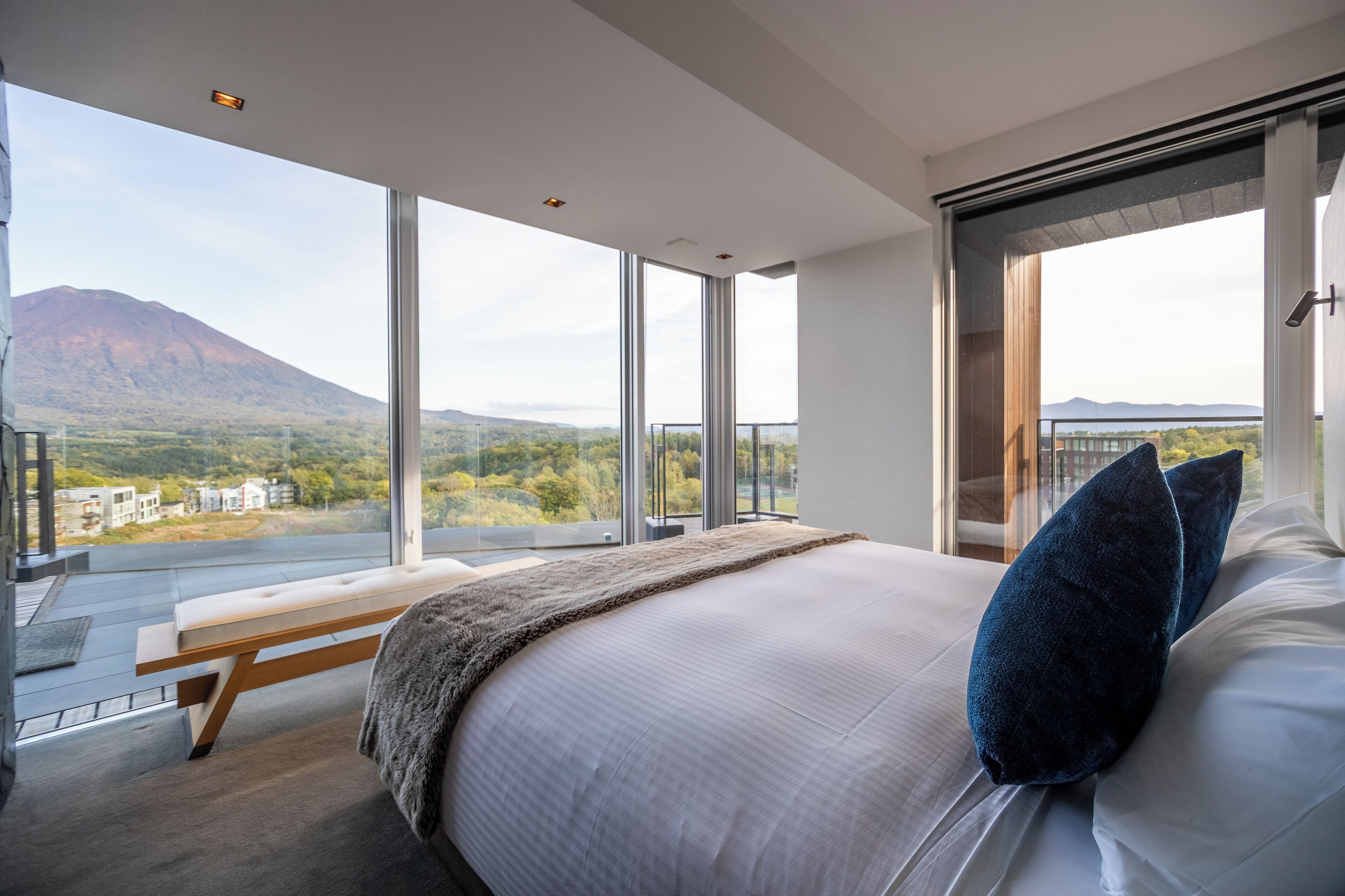 A modern bedroom with a large bed, a bench, and a view of a mountain and buildings.