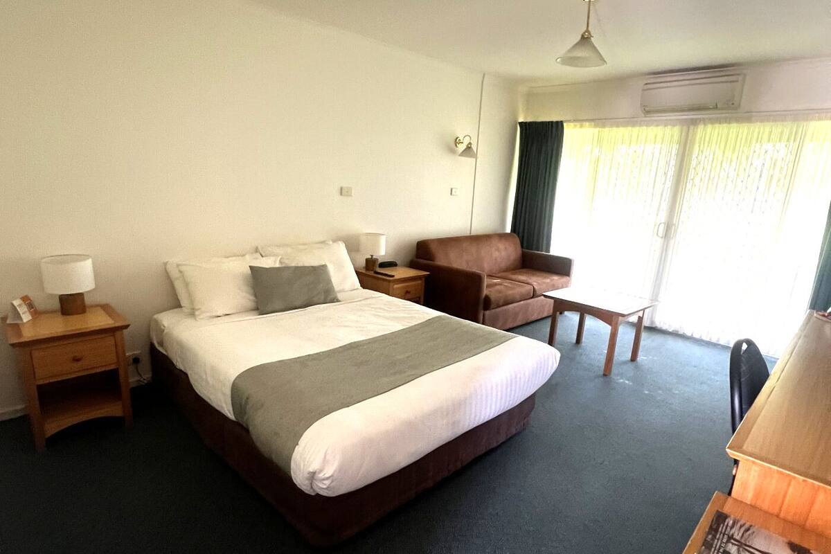 A hotel room with a large bed, a brown leather armchair, a small wooden table, and a desk with a lamp.