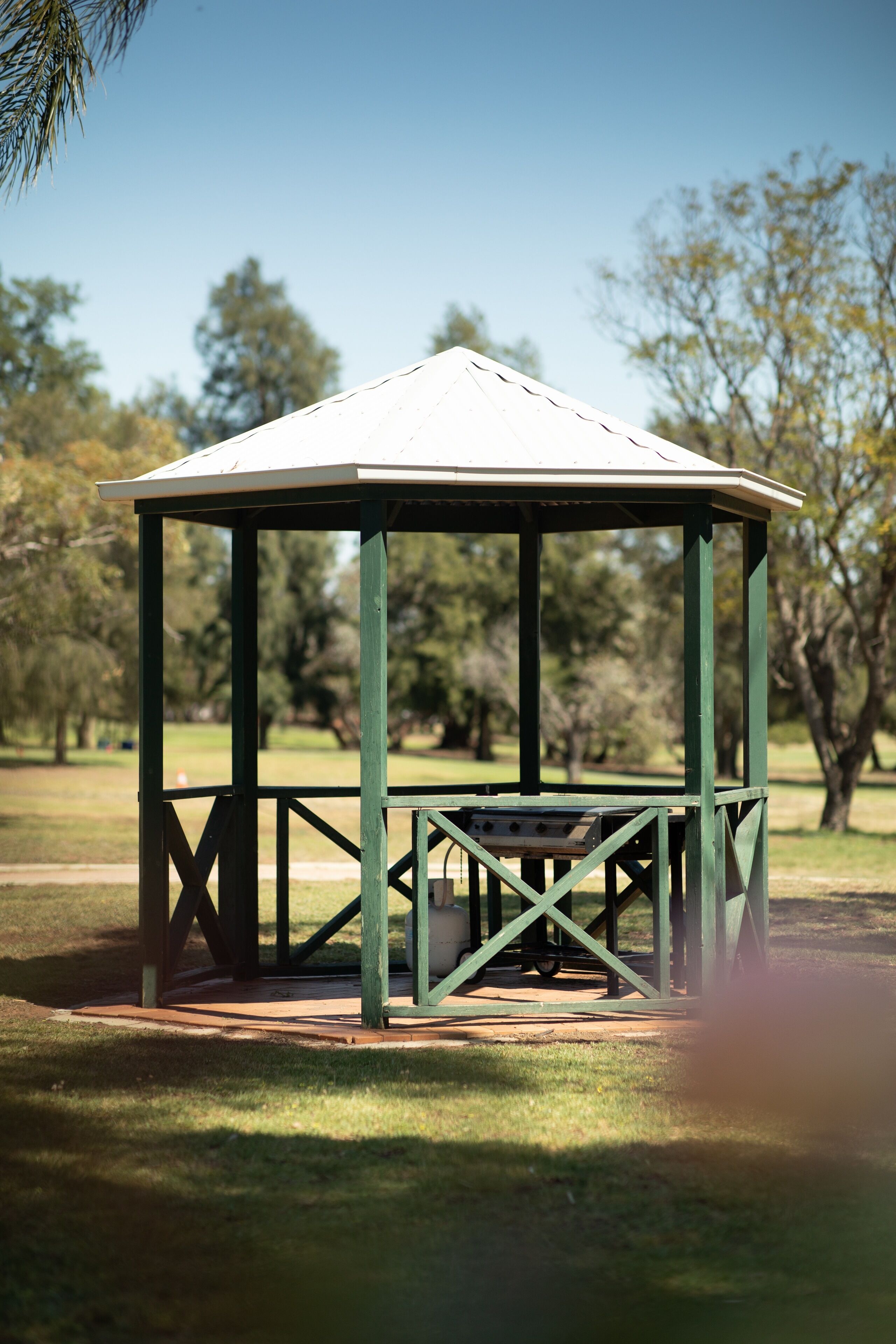 Un pabellón de diseño sencillo y abierto, ubicado en un parque con césped y árboles al fondo.
