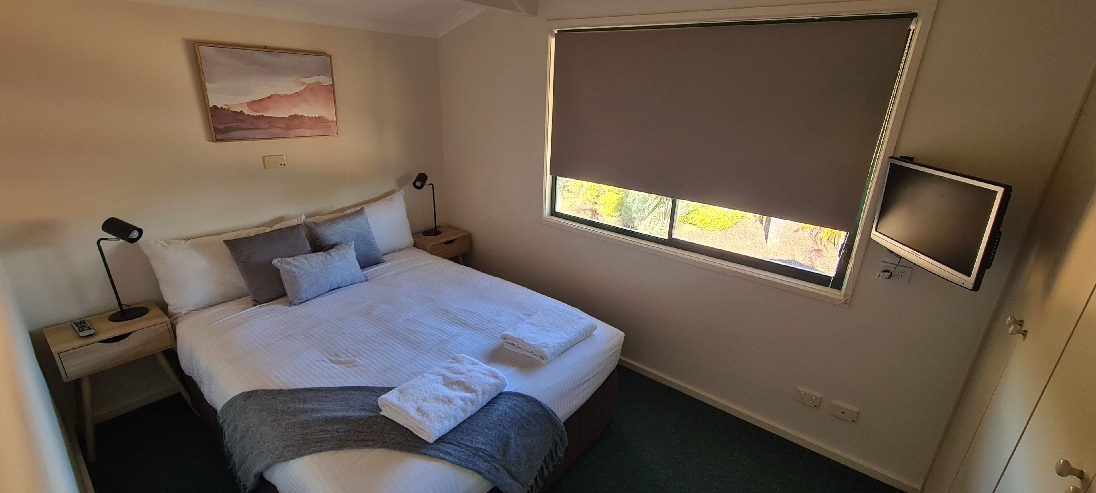 Un dormitorio con una cama grande, una mesita de noche con lámpara, un cuadro en la pared, una ventana con persiana y un televisor montado en la pared.
