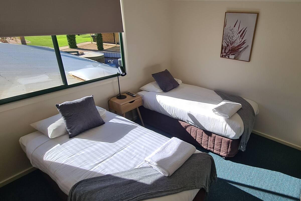 A small, clean room with two beds, a window, a wall-mounted TV, and a painting.