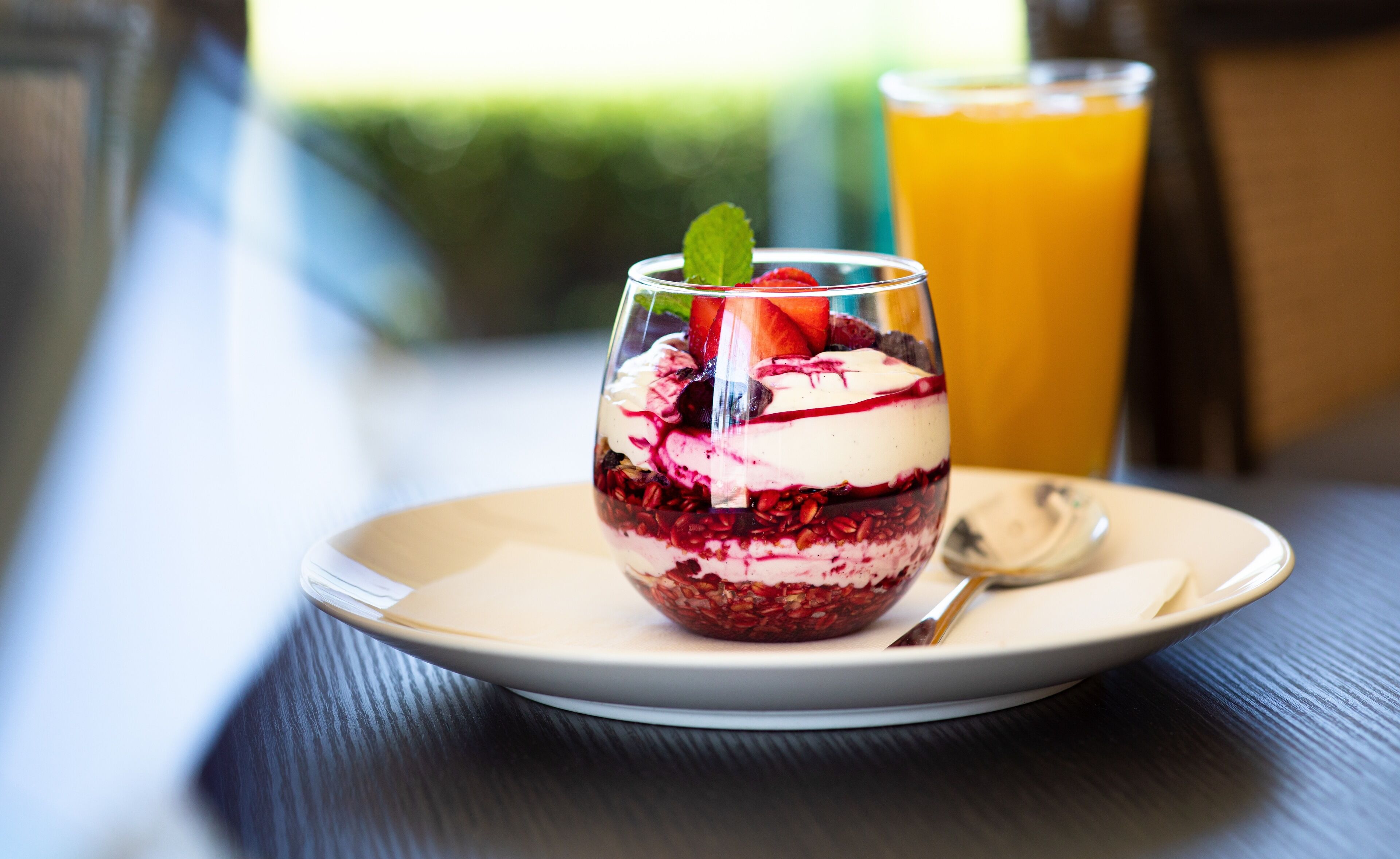 A dessert in a glass on a plate with a spoon and a glass of orange juice in the background.