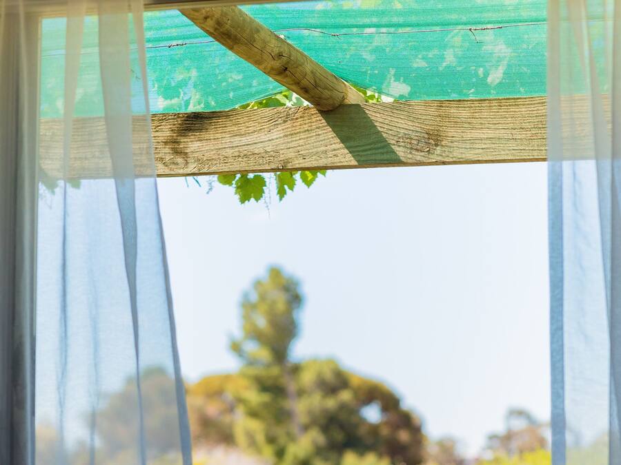 A patio with a table and chairs, a bottle, and a view of a garden through sheer curtains.