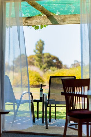 Une terrasse avec une table et des chaises, une bouteille, et une vue sur un jardin à travers de fines rideaux.