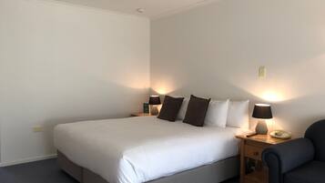 A neatly made bed with white bedding and brown pillows, a bedside table with a lamp, and a chair.