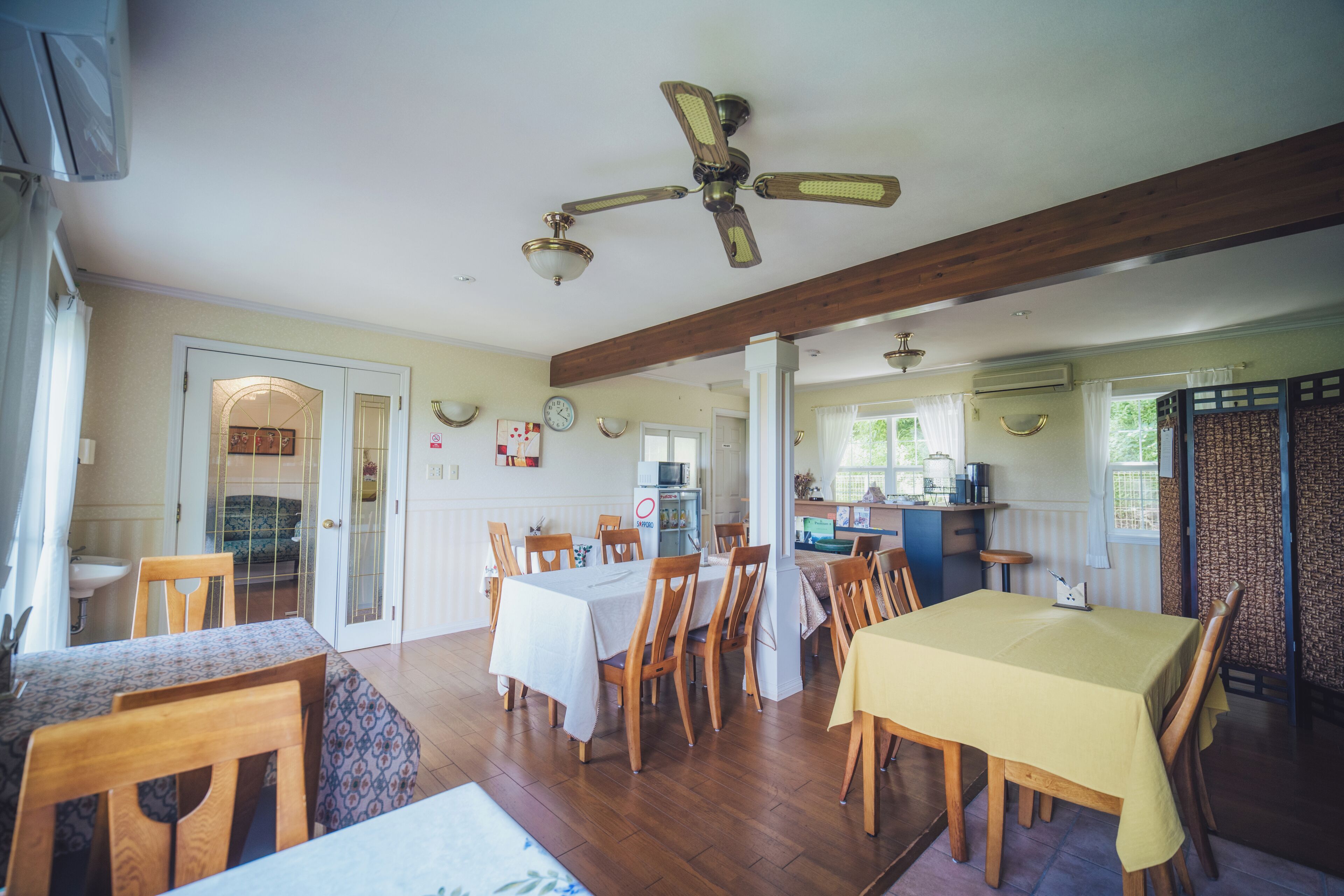 A dining area with wooden tables and chairs, a ceiling fan, and a kitchen in the background.