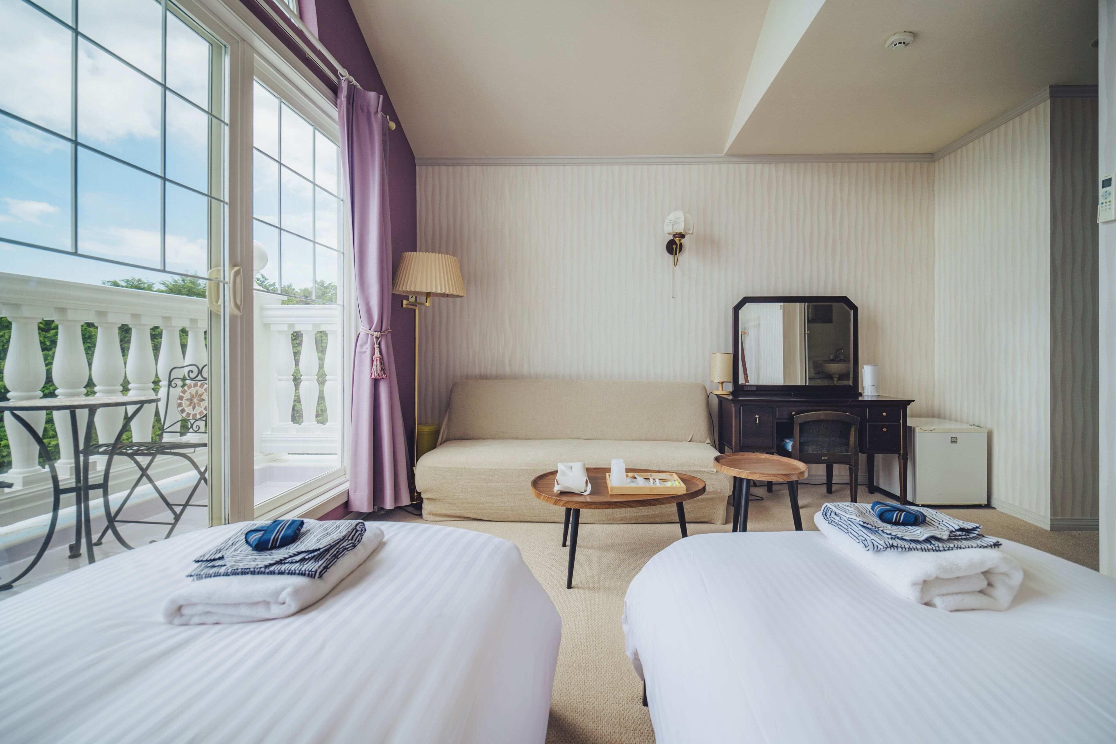 Ein Hotelzimmer mit zwei Betten, einem Schreibtisch, einem Sessel, einem kleinen Tisch und einem Balkon mit Tisch und Stühlen.