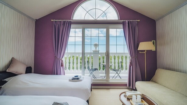 Habitación de hotel con dos camas, un sofá, un balcón con mesa y sillas, y vista al cielo.