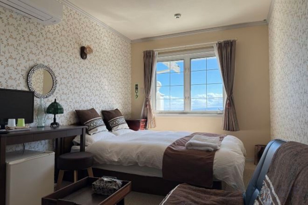 Una habitación de hotel con cama, escritorio, silla, ventana con cortinas y un espejo montado en la pared.