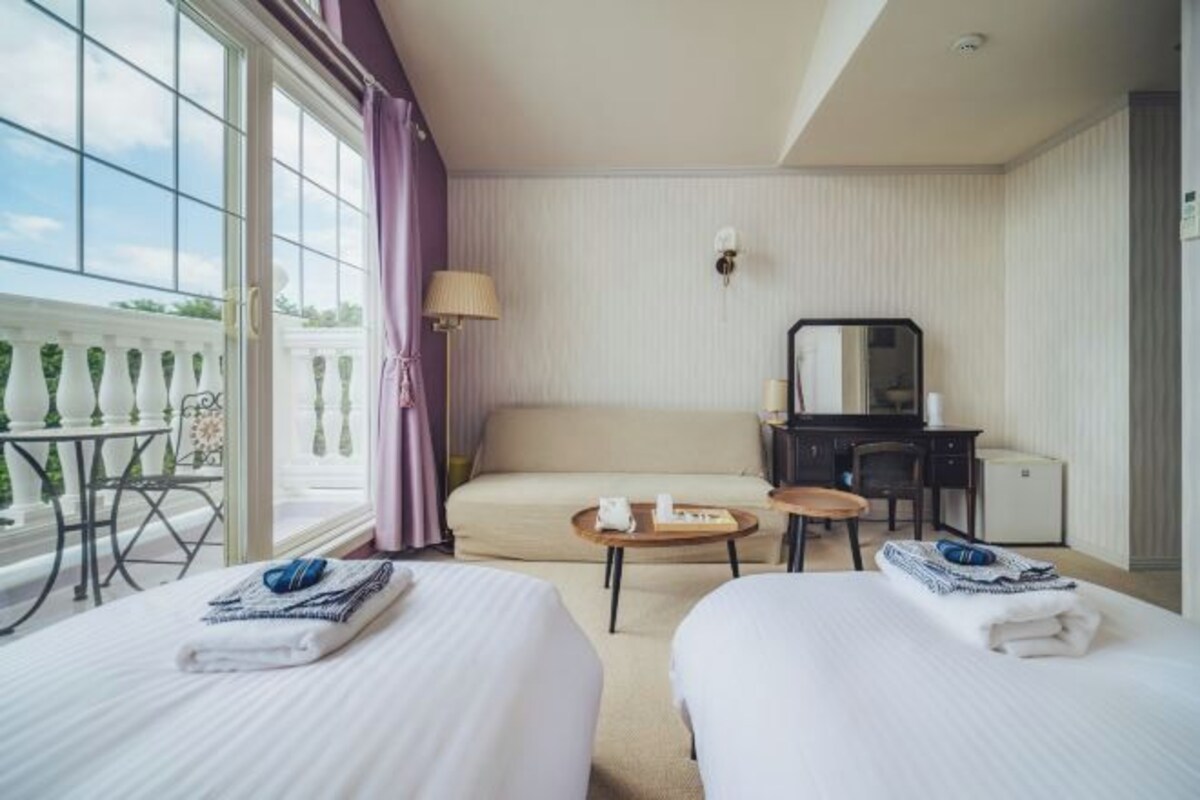 Habitación de hotel con dos camas, un escritorio, una silla, una mesita y un balcón con mesa y sillas.