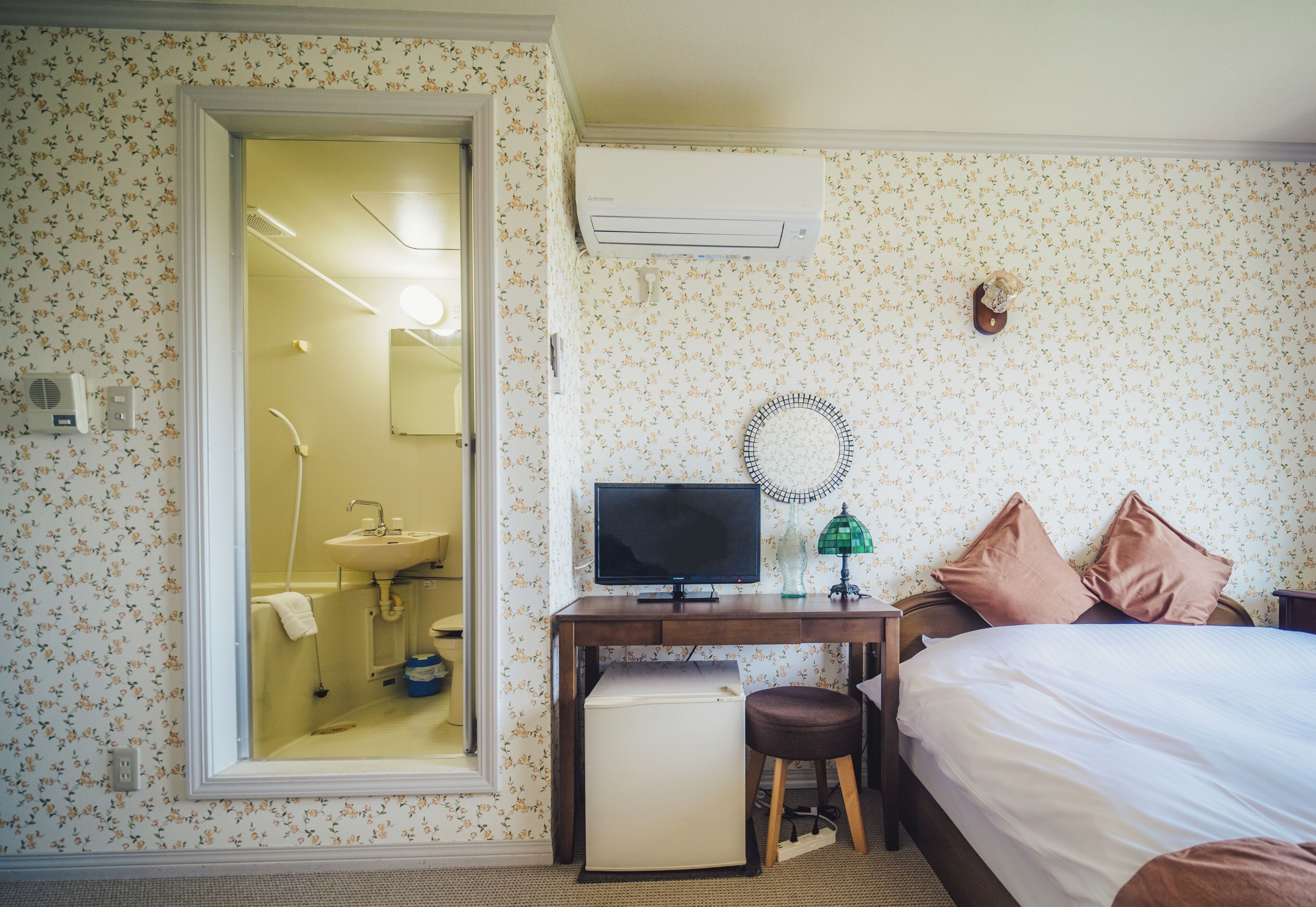 A hotel room with a bed, a desk with a computer, a small refrigerator, a mirror, and an air conditioning unit.