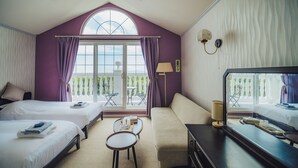 Habitación de hotel con dos camas, un sofá, una mesita, una lámpara y un ventanal grande con cortinas.