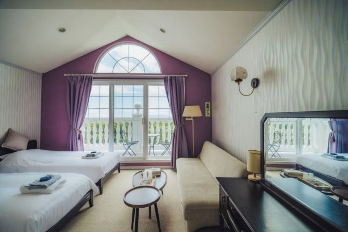 A hotel room with two beds, a sofa, a small table, a TV, and a balcony with a view.