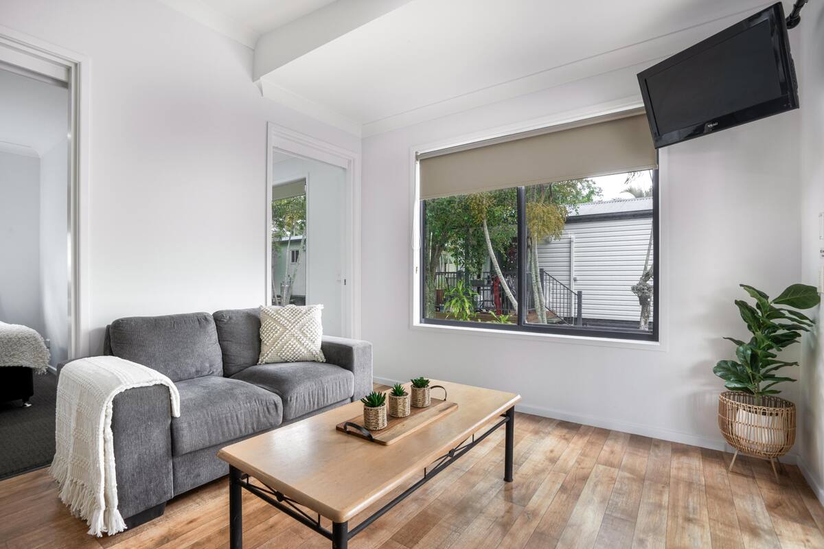 Una sala de estar moderna con un sofá gris, una mesa de centro de madera y un ventanal con vistas a una casa y árboles.