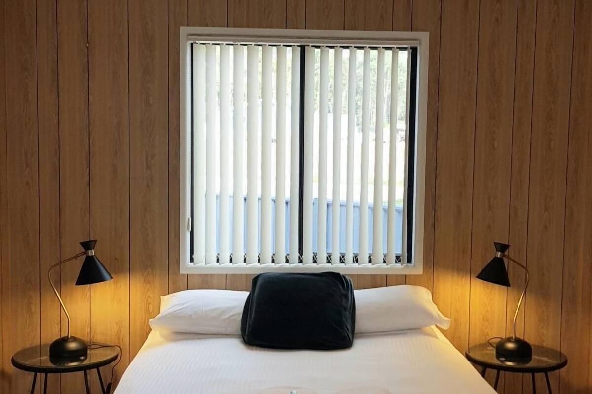 Una cama impecablemente hecha con sábanas blancas y un almohadón negro, flanqueada por dos mesitas de noche negras con lámparas, en una habitación con paneles de madera y un ventanal con persianas verticales.