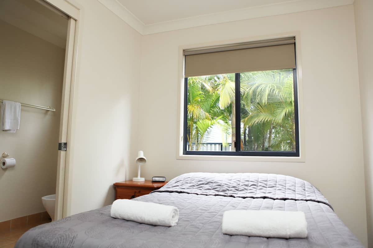 Un dormitorio con cama, mesita de noche, ventana con vistas a palmeras y puerta que da al baño.